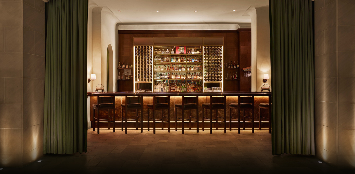 A dimly lit bar with a long counter and stools, shelves of bottles in the background.